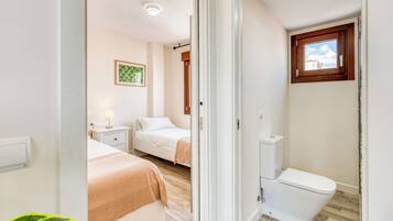 A bedroom with two beds, a bedside table with a lamp, a window with a view, and a white toilet.