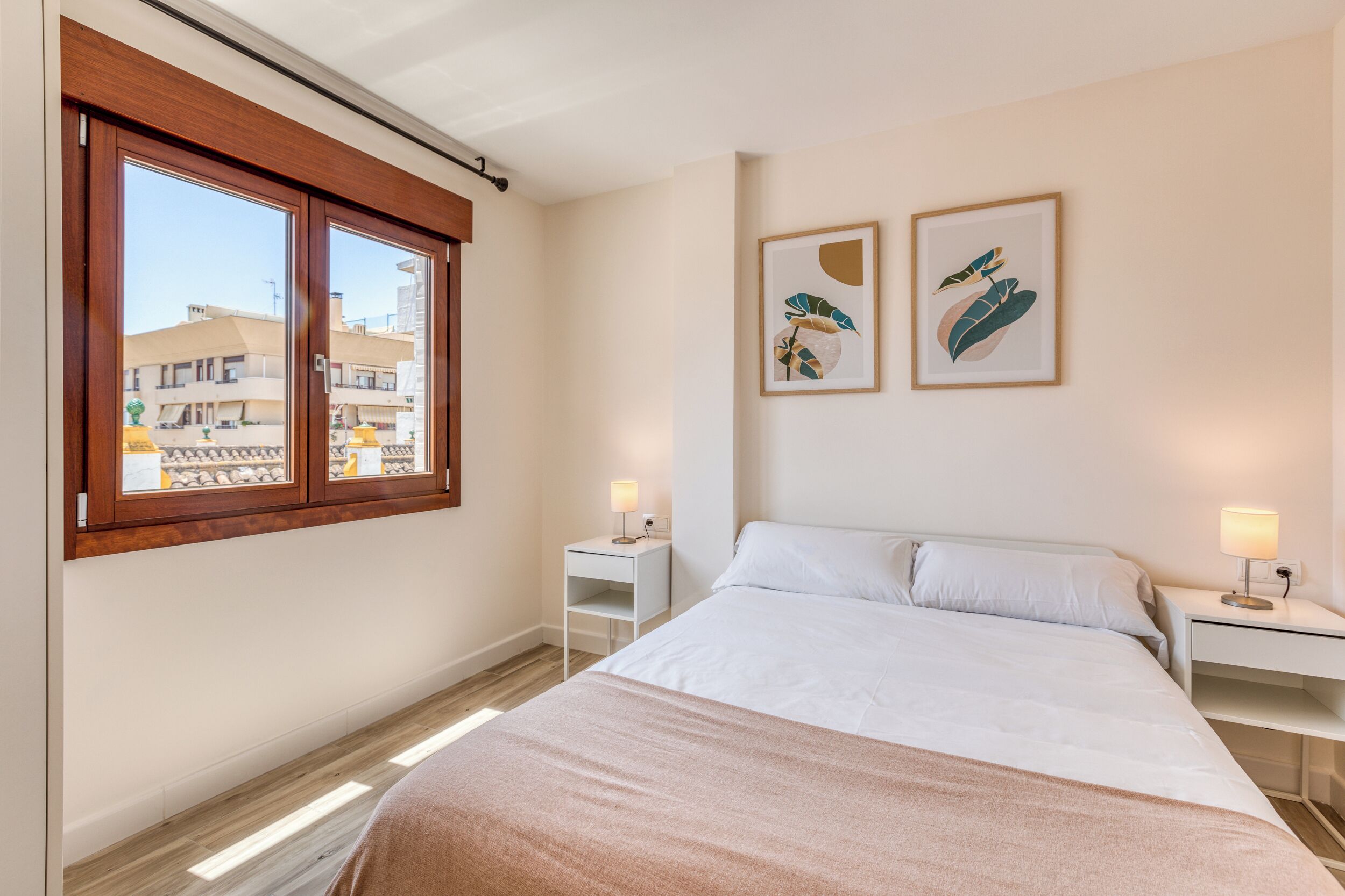 Ein Schlafzimmer mit einem Bett, zwei Nachttischen mit Lampen, einem Fenster mit Jalousien und zwei gerahmten Wandbildern.