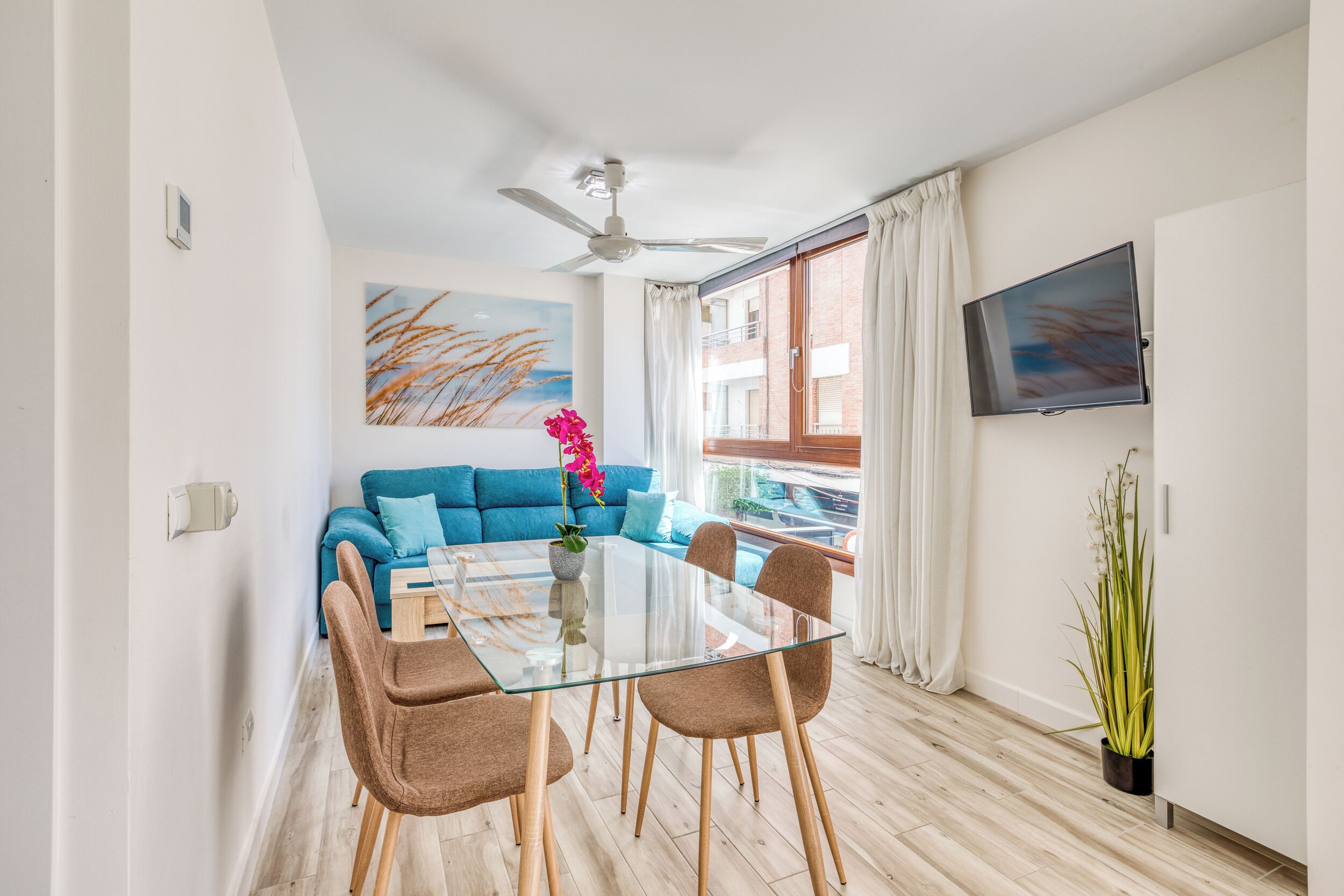 A modern living room with a glass dining table, blue sofa, and a ceiling fan.