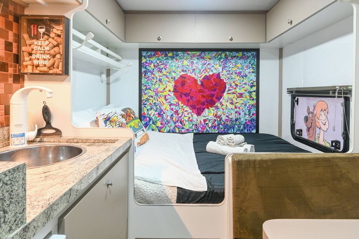 Une chambre compacte et moderne, dotée d’une kitchenette, d’un lit avec un mur décoré d’un motif en forme de cœur aux couleurs vives, et d’un petit téléviseur.