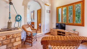 Un salón acogedor con muebles de mimbre, una chimenea de piedra, un televisor y ventanas amplias con vista a árboles.