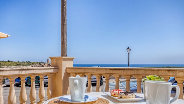 Una mesa con una taza y platillo blancos, un plato con un postre, y una taza blanca en un balcón con vista al mar.