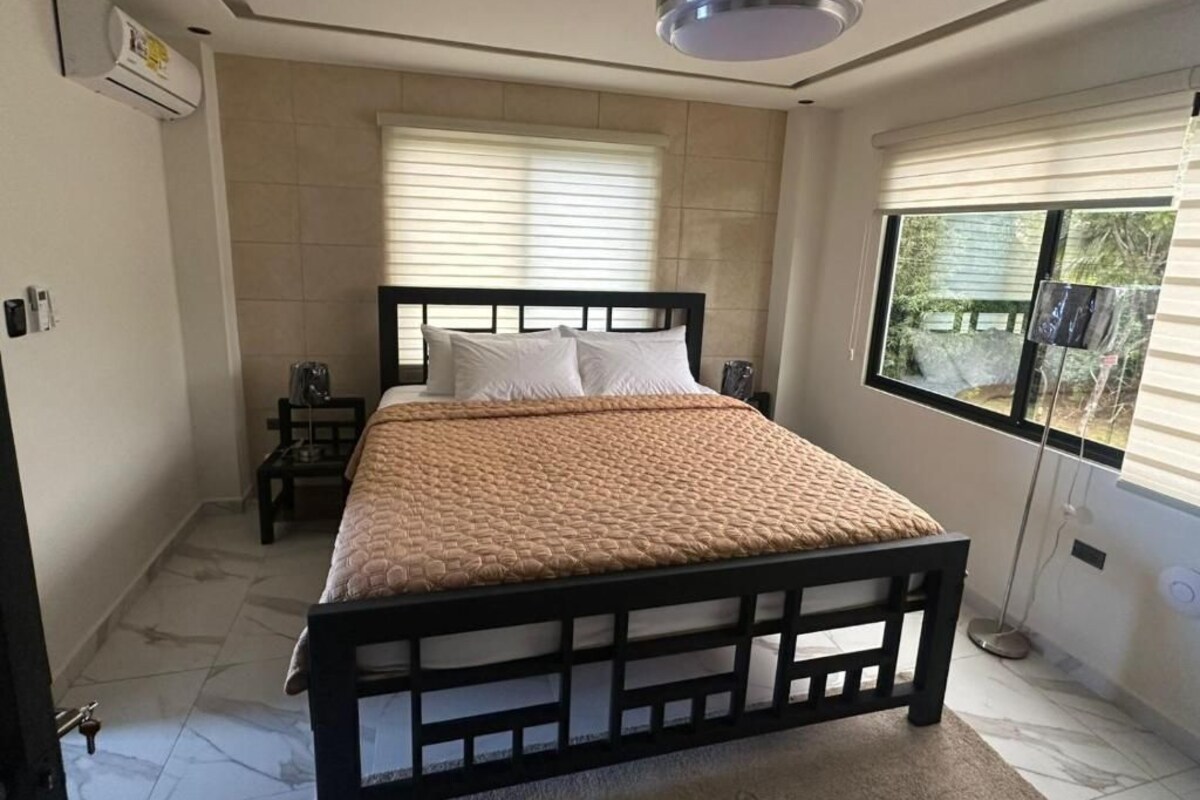 Ein Schlafzimmer mit Bett, Deckenventilator, Fenster mit Jalousien und Blick ins Freie.