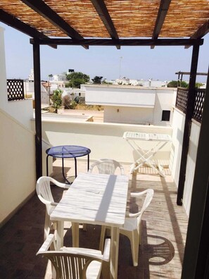 A covered outdoor seating area with a table and chairs, a small round table, and a view of the surrounding buildings. - Apartment just 200 m from the beautiful beach. Private parking. (Torre San Giovanni)