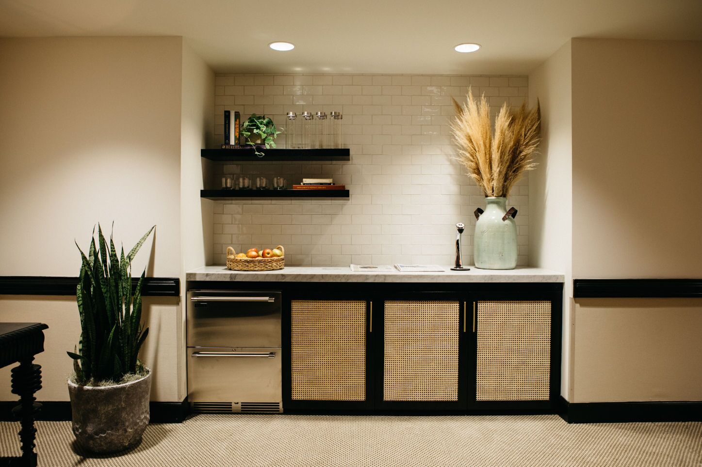 A kitchen area with a white tiled backsplash, a black countertop, and a basket of fruits. There are two wicker baskets below the countertop, a potted plant on the left, and a vase with dried plants on the right.