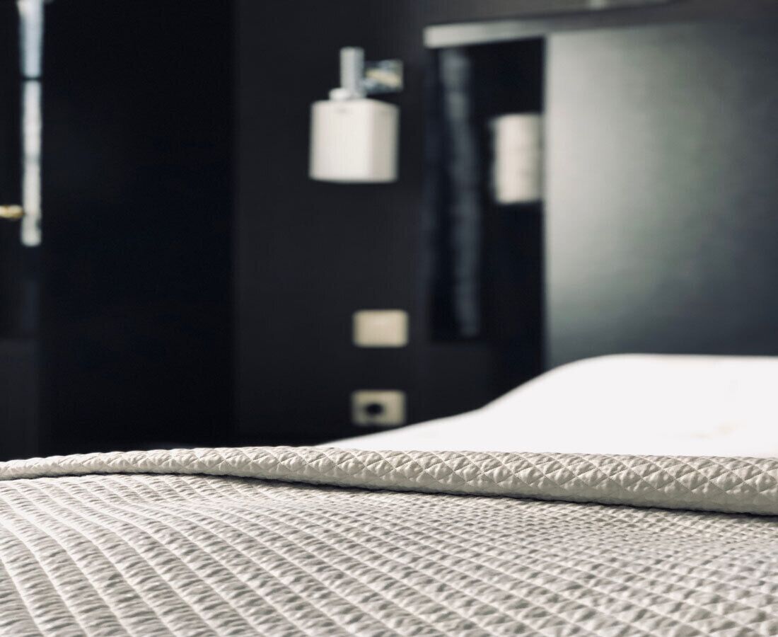 A close-up of a bed with a textured white cover, a dark wall with a white object on it, and a black object on the wall.