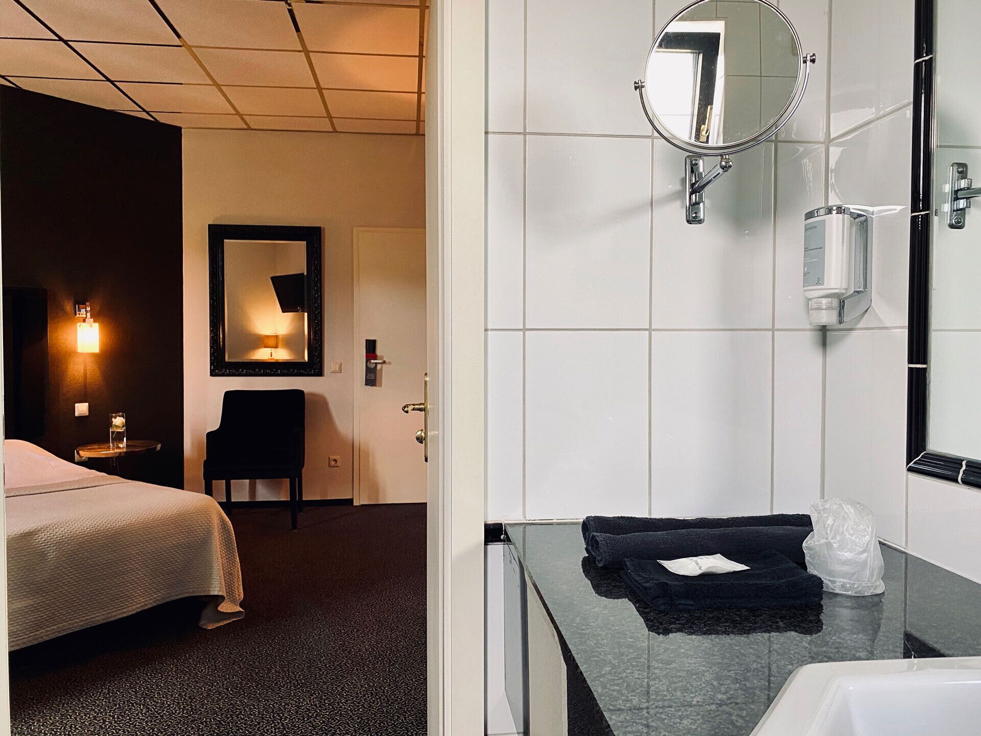 Ein Hotelzimmer mit einem Bett, einem Sessel, einem Schreibtisch, einem Spiegel und einem Badezimmer mit Waschbecken und Handtuch.