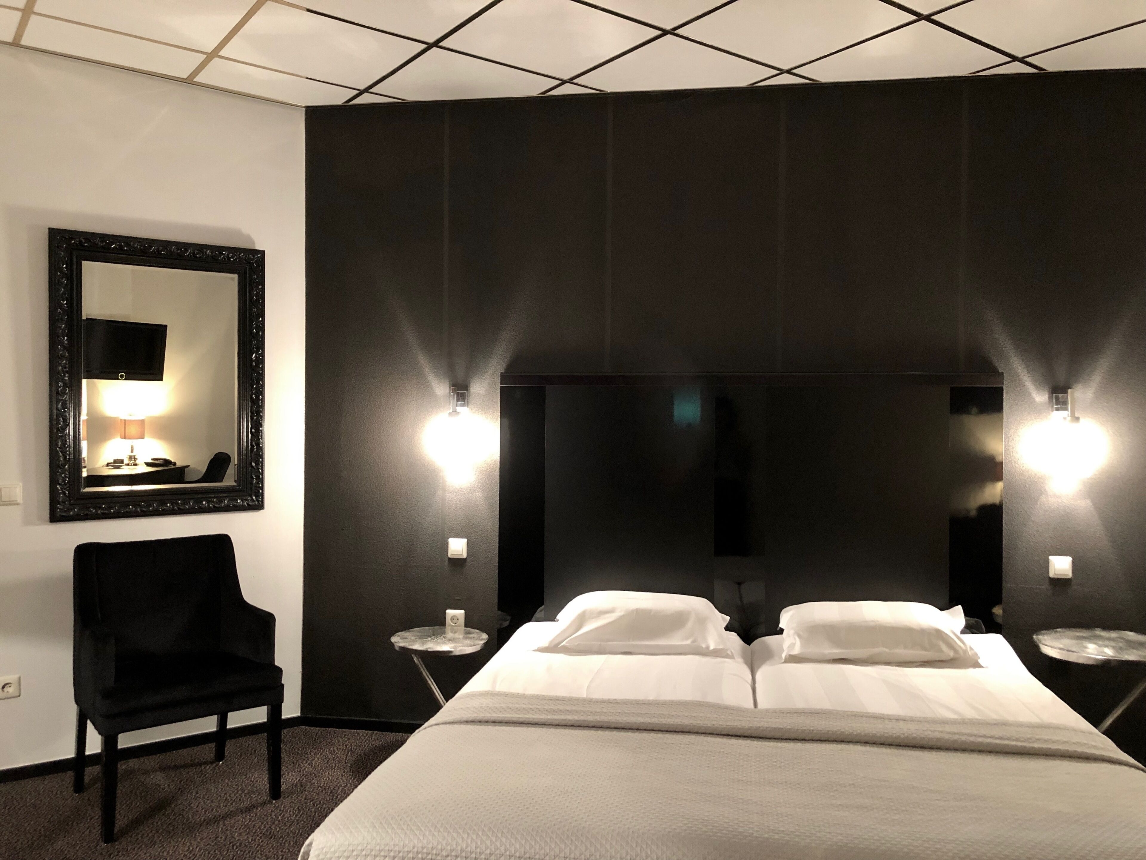 Ein Hotelzimmer mit einem großen Bett, zwei Nachttischen, einem Sessel, einem Spiegel und Wandleuchten.