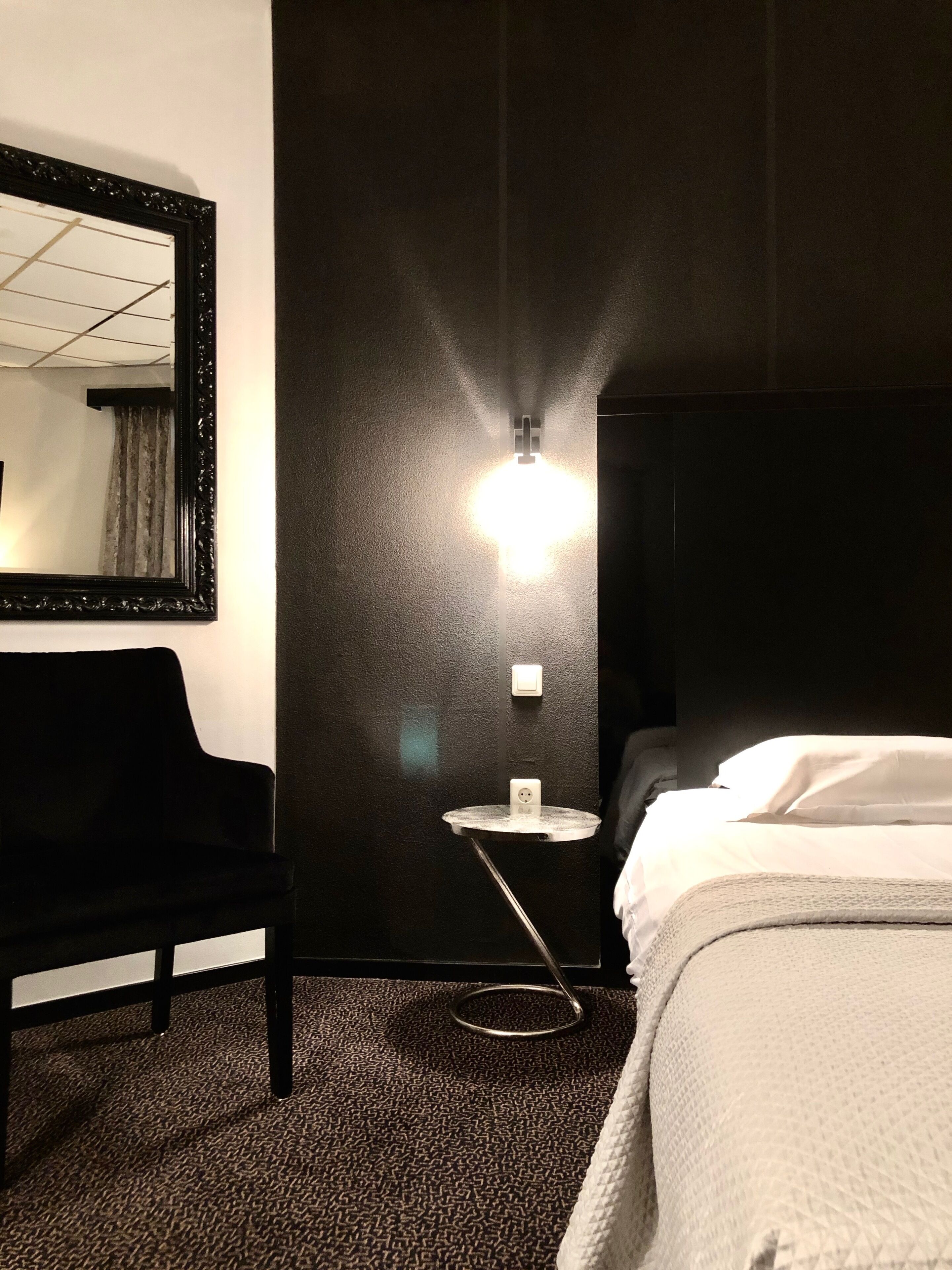 A hotel room with a black wall, a round side table with a lamp, a black chair, and a bed with white bedding.