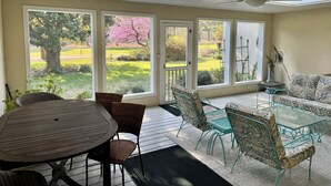 A covered patio with a table and chairs, large windows with a view of a garden and trees, and a ceiling fan. - North End Villa-behind the Cavalier2blocks2 beach (Virginia Beach)