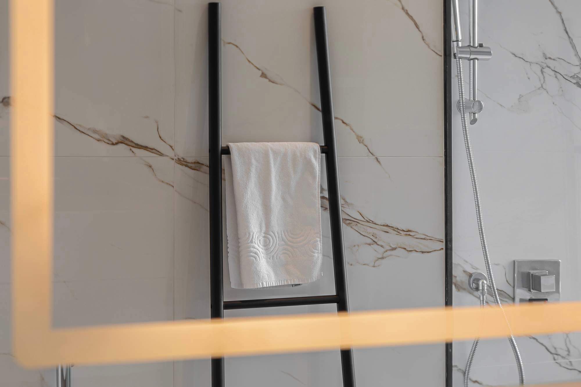 Une salle de bain avec un mur en marbre, une serviette suspendue à un porte-serviettes noir et une douche équipée d’un pommeau de douche à main.