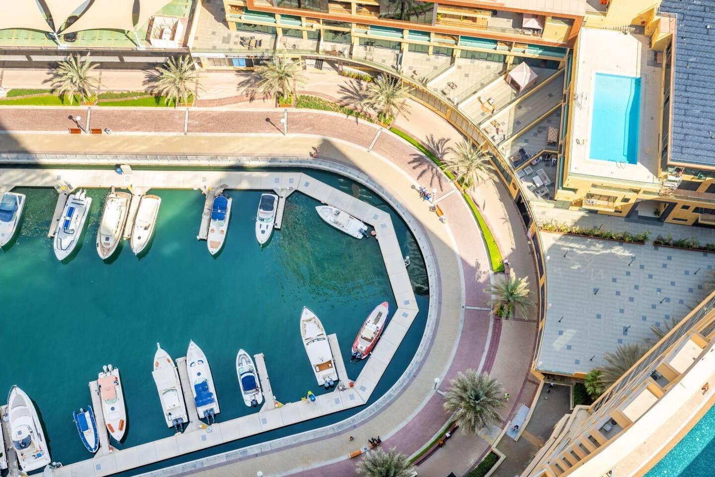 An aerial view of a marina with boats docked in a circular arrangement, surrounded by palm trees and a modern building with a pool.
