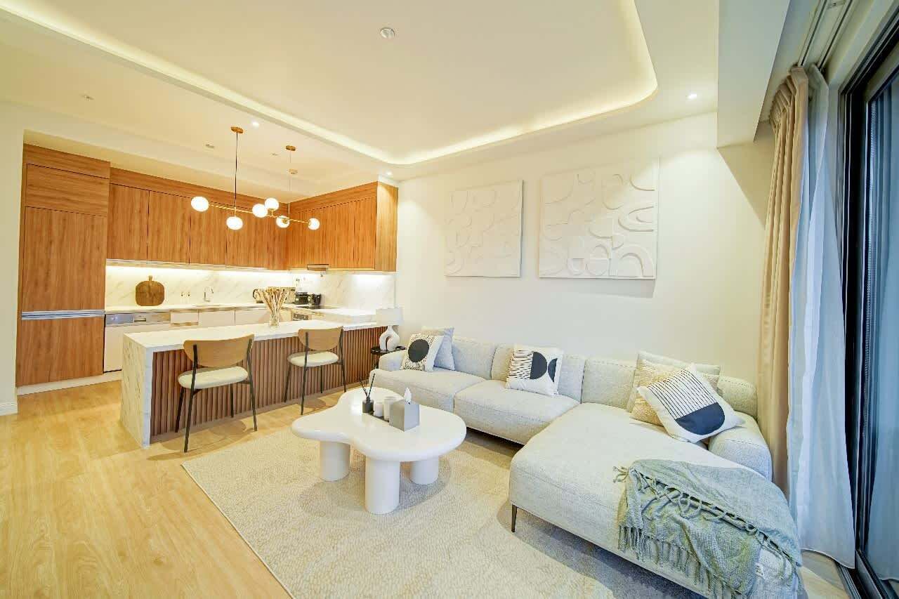 A modern living room with a sofa, coffee table, and chairs. There is a kitchen area with wooden cabinets and a dining table.