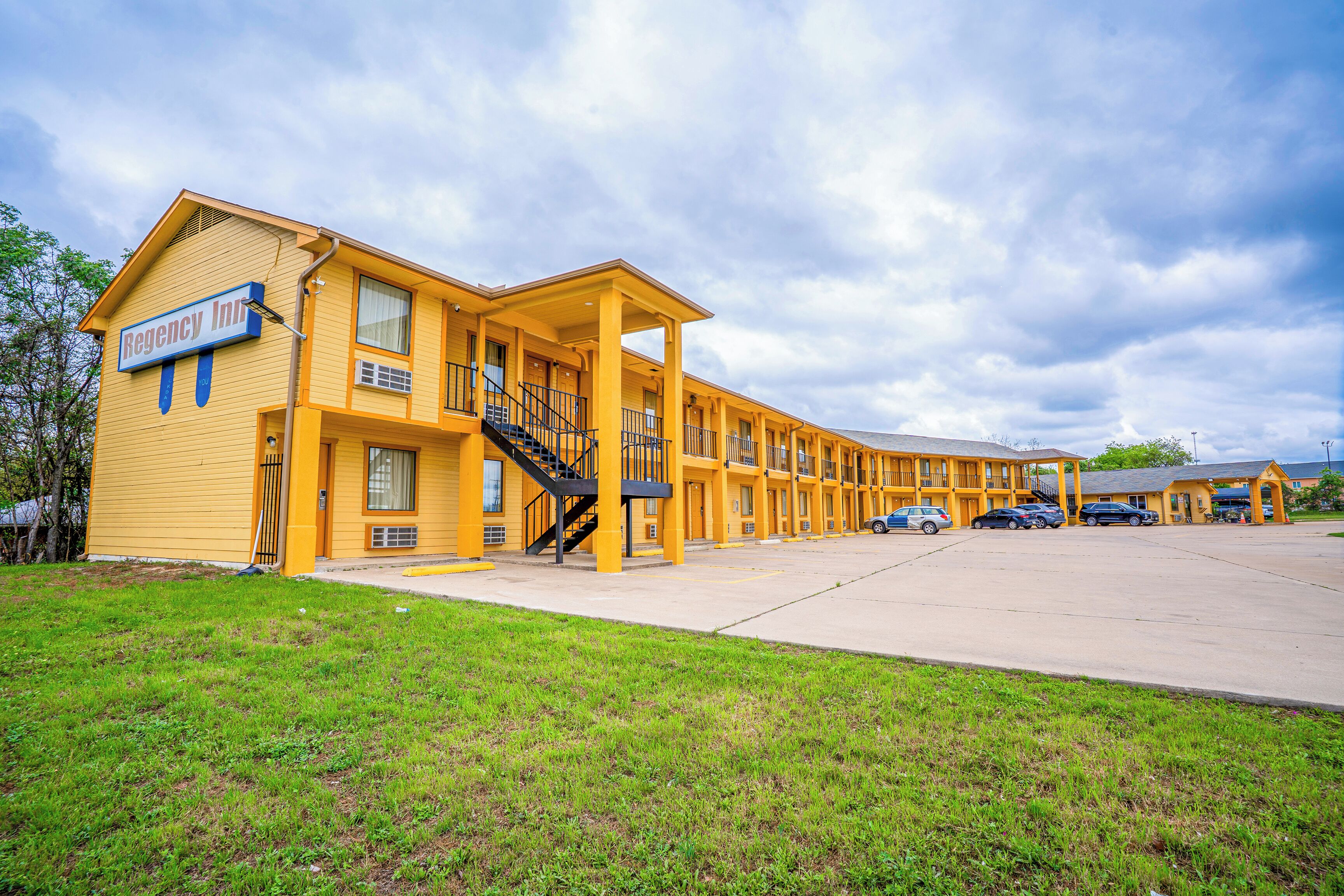Un motel amarillo con un letrero que dice "Regency Inn", que cuenta con una escalera central, varios balcones y un estacionamiento.