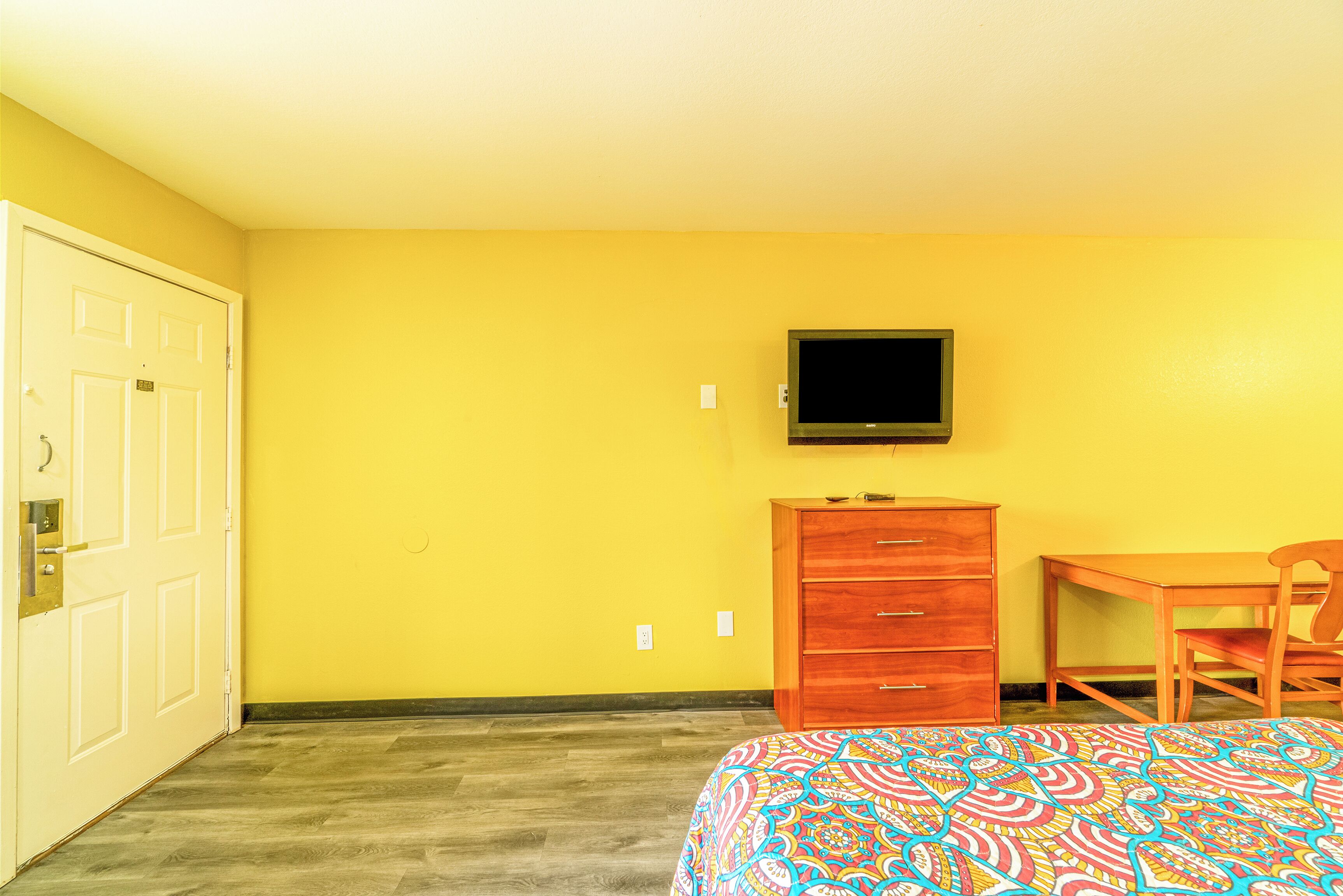 Una habitación con una cama, un aparador de madera, un televisor montado en la pared y una puerta.