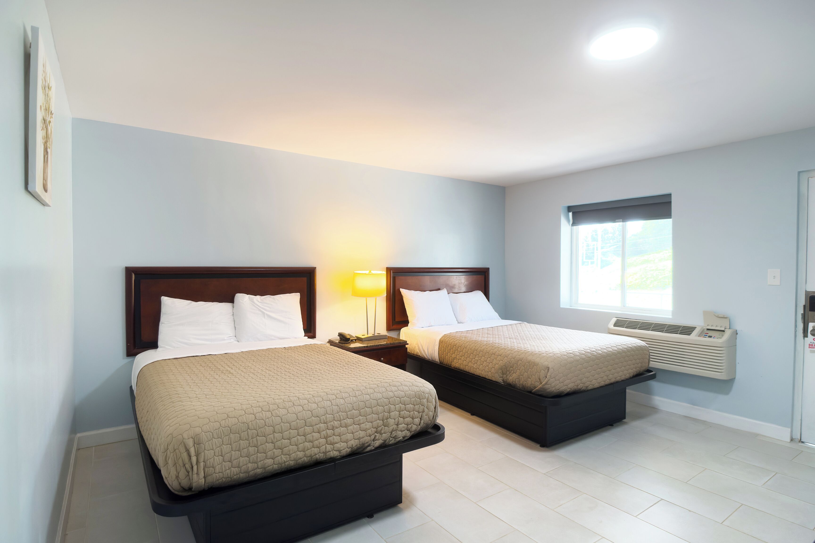 A hotel room with two beds, a nightstand with a lamp, a window with an air conditioner, and a door with a lock.