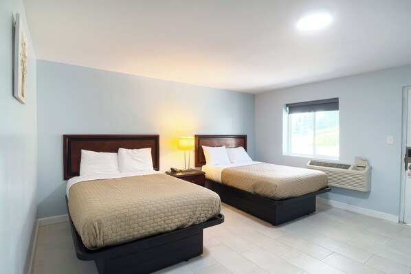 A hotel room with two beds, a nightstand with a lamp, a window with an air conditioner, and a door with a lock.