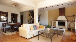 A spacious living area with a sofa, coffee table, and dining table. A fireplace and staircase are visible in the background. - House Sea View with Communal Pool (Corfu)