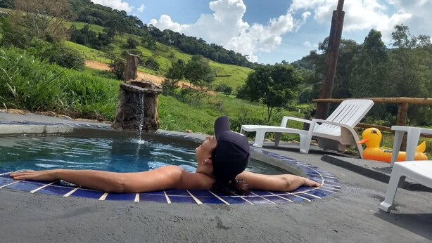 A person lying on a blue mat in a small pool with a waterfall feature, surrounded by greenery and white lounge chairs.