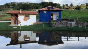 A small blue building with a red roof, a brick structure with a white wall, and a pond reflecting the buildings.
