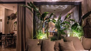 Un salon moderne avec un canapé, de grandes plantes vertes et un coin repas visible à travers une cloison en verre.