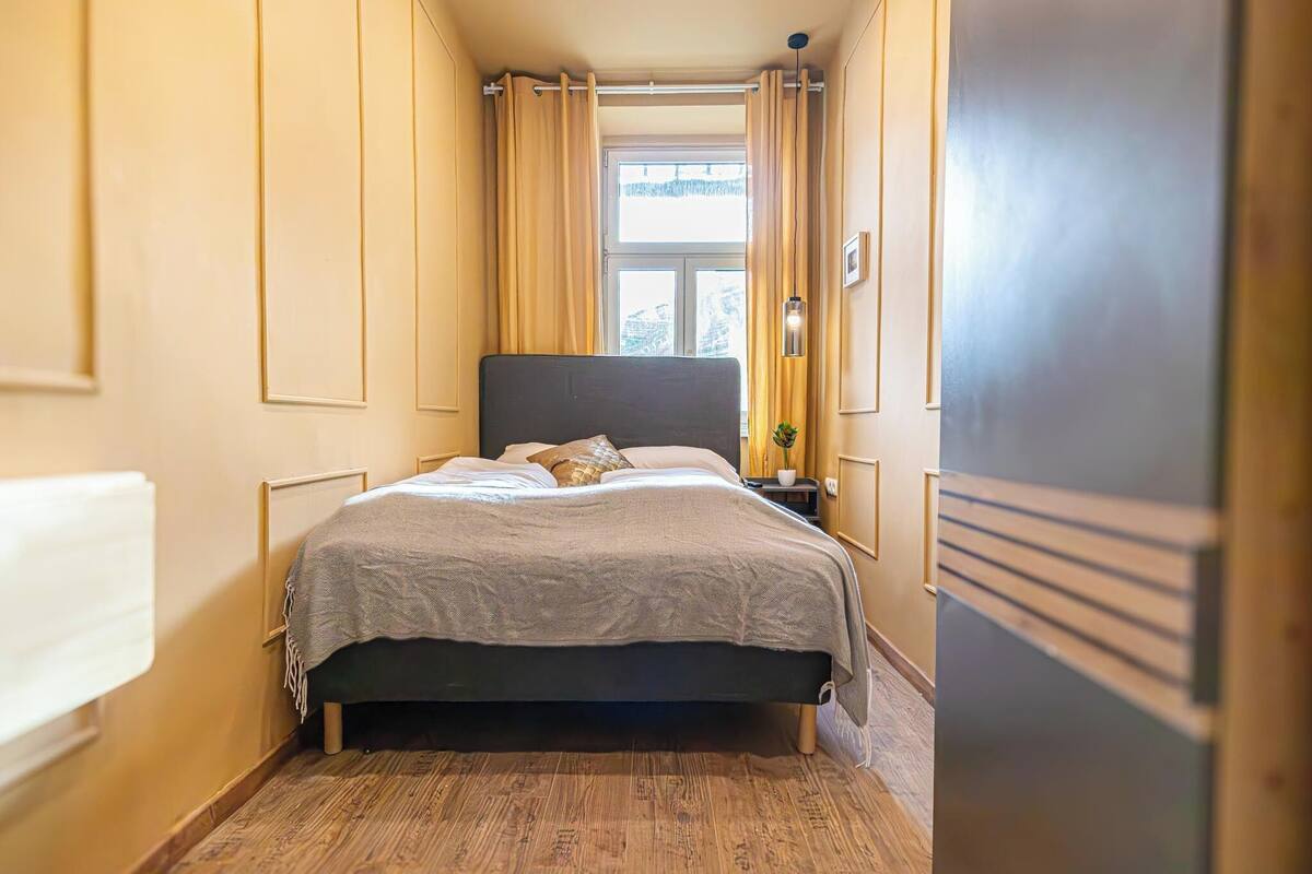 A narrow hotel room with a bed, a nightstand, a window with curtains, and a wooden floor.