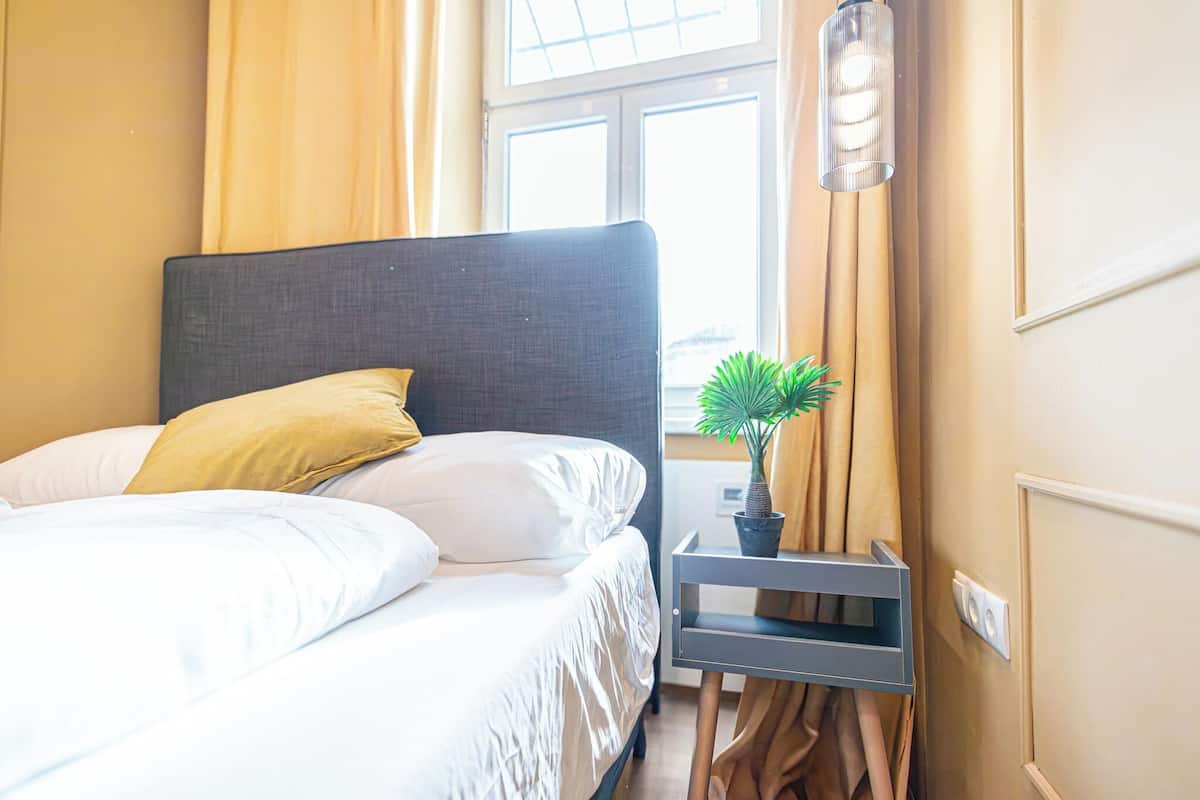 A neatly made bed with white linens and a single yellow pillow, a small potted plant on a bedside table, and a window with curtains.