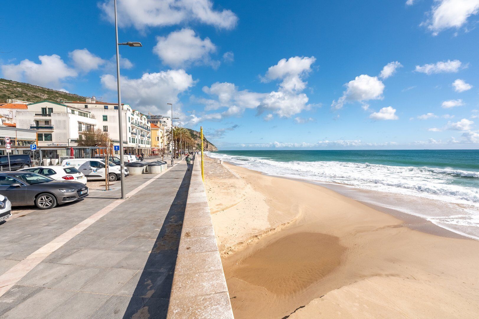 Eine Küstenstraße mit parkenden Autos, einem Fußweg und Gebäuden mit Balkonen mit Blick auf den Strand.