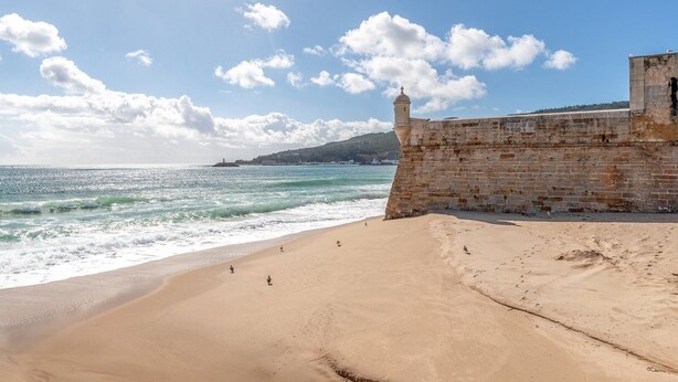 Una playa con un muro de piedra y un faro al fondo.