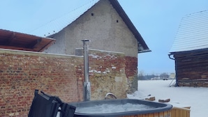 Een houten jacuzzi in een besneeuwde omgeving, met een bakstenen muur en een houten gebouw op de achtergrond.