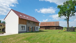 Een modern huis met een schuin dak, een grote boom in de voortuin en een schuur op de achtergrond.