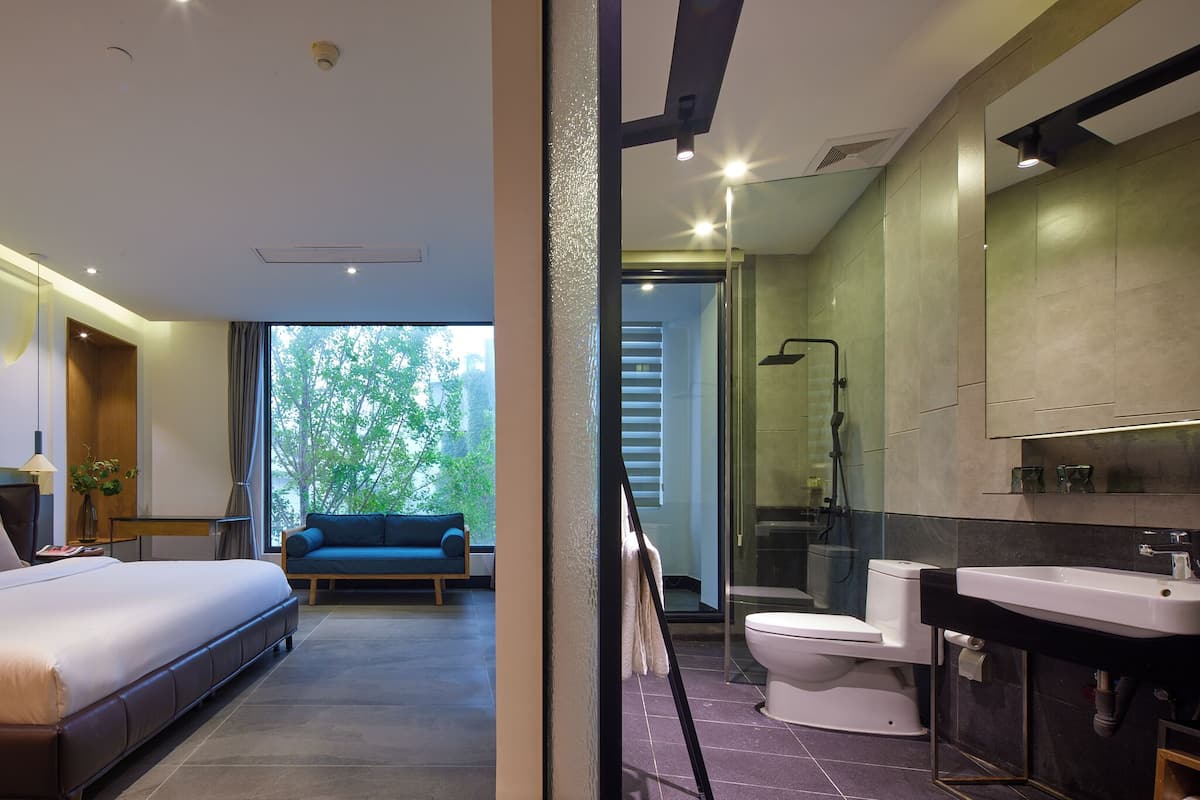 A modern hotel room with a large bed, a sofa, a bathroom with a shower, and a view of trees.