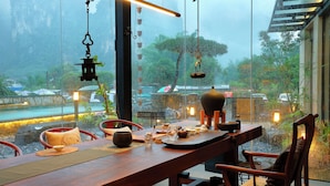 A dining area with a wooden table set for two, a hanging lantern, and a view of a rainy outdoor scene through large windows. - Yangshuo The One Hotel (Guilin)
