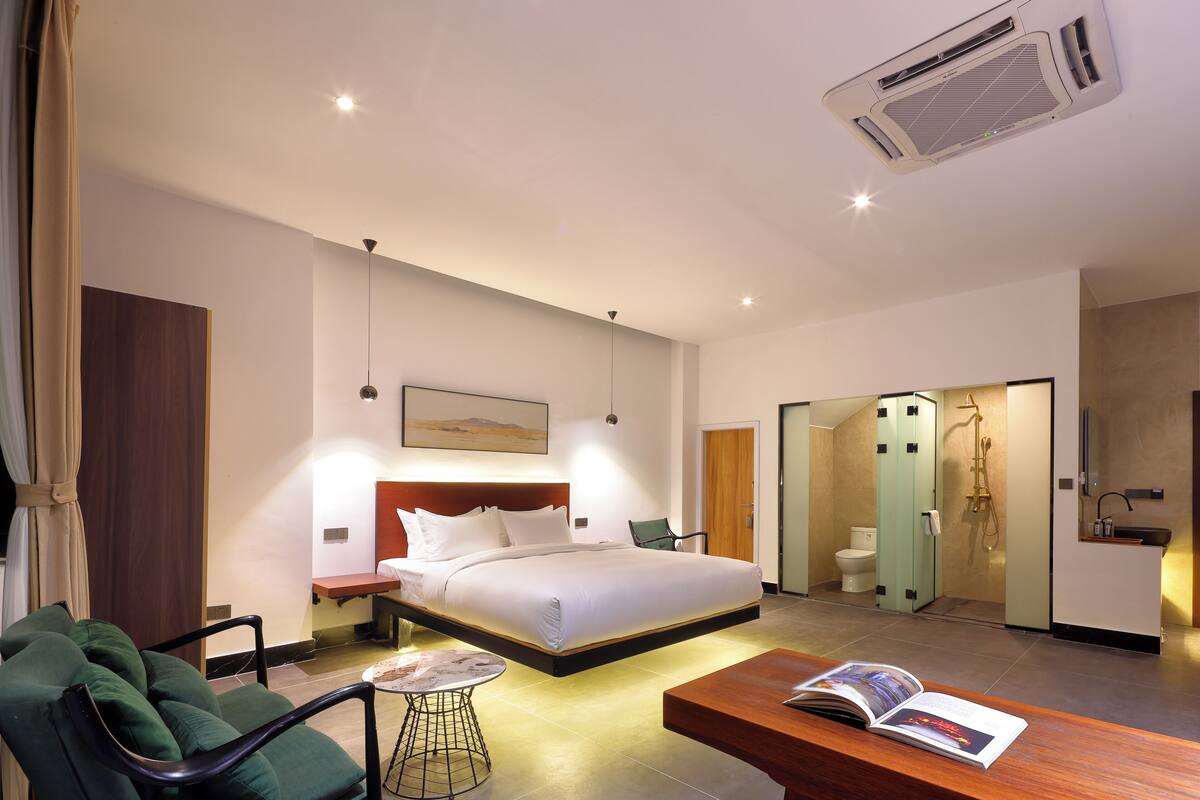 A modern hotel room with a large bed, a desk, a chair, and a small table. There is a bathroom with a shower and toilet visible through an open door.