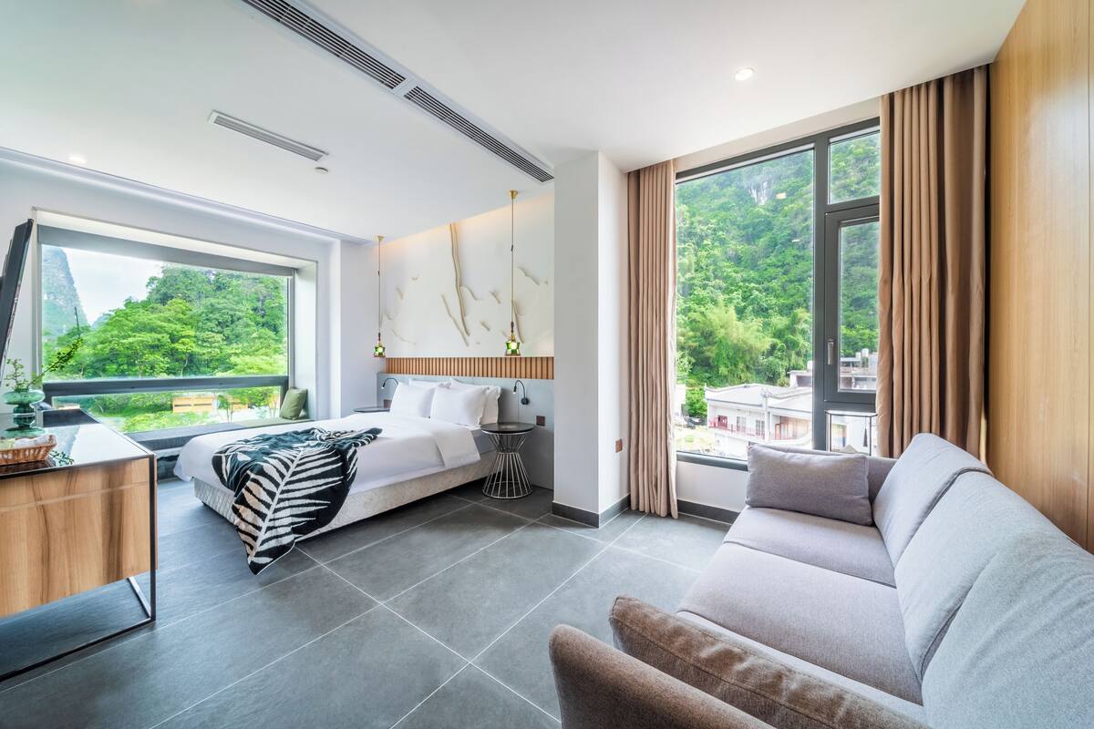 A modern hotel room with a bed, a sofa, a desk, and a large window offering a view of greenery.