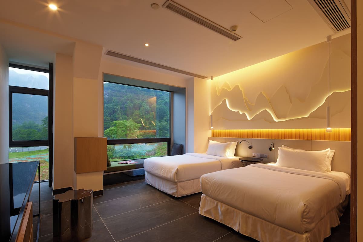 A modern hotel room with two beds, a large window with a view of greenery, and a unique wall design.