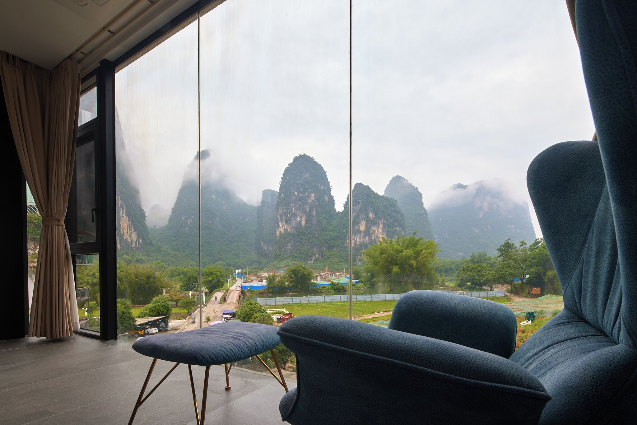 A room with a large window offering a view of mountainous terrain and a river. There is a blue sofa and a small table in the foreground.