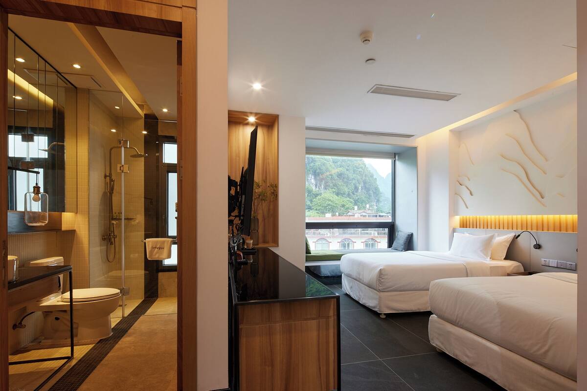 A modern hotel room with a glass shower, a wooden vanity, two beds, and a view of a bridge.