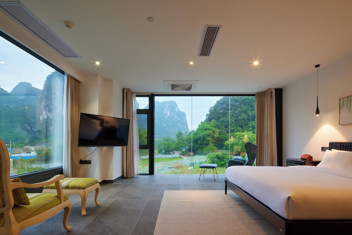 A hotel room with a large bed, a TV, a chair, and a view of a mountainous landscape through a glass door.