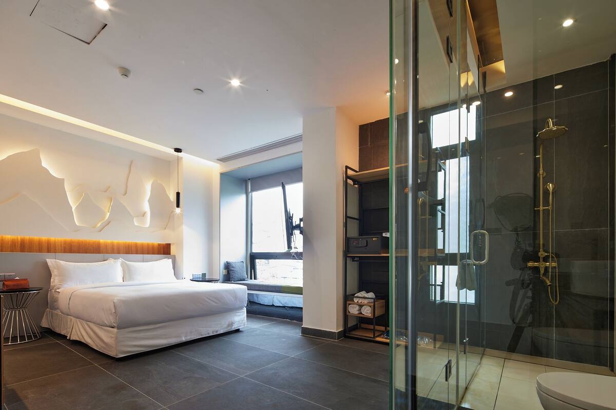 A modern hotel room with a large bed, a glass-enclosed shower, and a view of the outdoors.