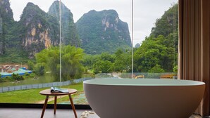 A modern bathroom with a large bathtub, a small table, and a view of a river and mountains. - Yangshuo The One Hotel (Guilin)