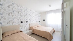 A hotel room with two beds, a small bedside table, a wall-mounted light, and a window with sheer curtains. - Newly Renovated Modern two Room Apartment (San Michele al Tagliamento)