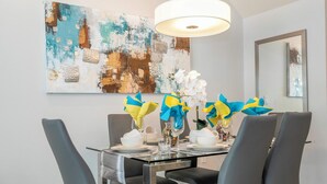 Un coin repas avec une table en verre, quatre chaises, un lustre moderne et un grand tableau abstrait accroché au mur.