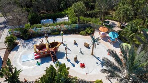 Vista aerea di un parco con un'area giochi a tema piratesco, palme e una zona recintata con posti a sedere all'aperto.