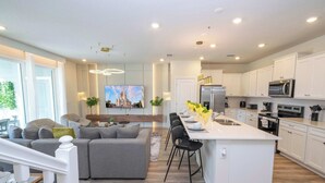 A modern kitchen with white cabinets, a central island, and a dining area with a grey sofa and black chairs. - Fabulous 5Bd Pool & Game Room @ Windsor Cay 1253 (Clermont)