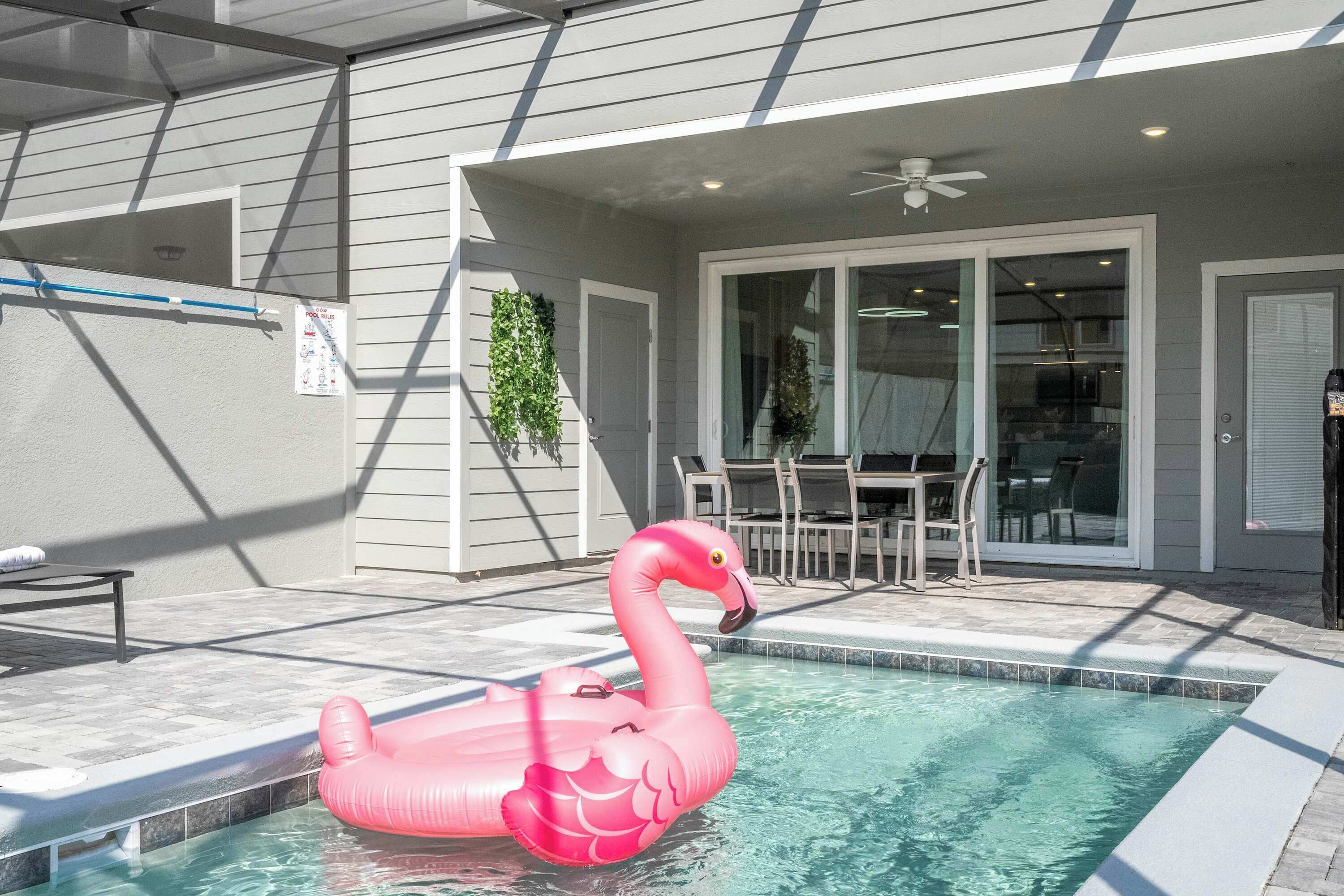A swimming pool with a pink flamingo float, a covered patio area, and a dining table with chairs.