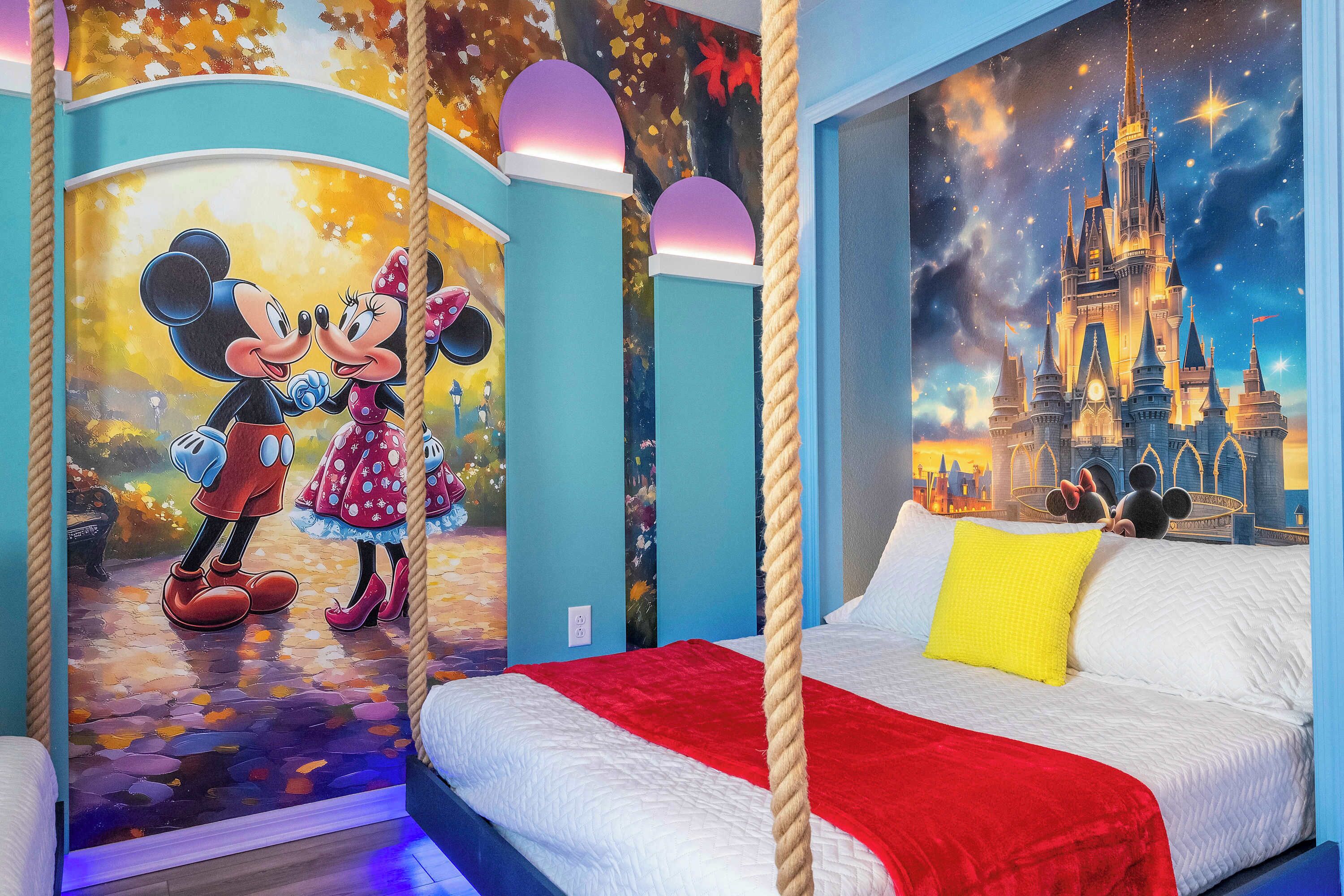 Une chambre avec une fresque murale représentant Mickey et Minnie Mouse, une autre fresque représentant un château, un lit avec une literie blanche et un plaid rouge, et une barrière de lit en corde.