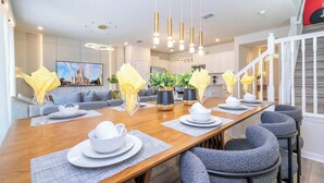 A modern dining area with a long wooden table set for a meal, surrounded by grey upholstered chairs. - Fabulous 5Bd Pool & Game Room @ Windsor Cay 1253 (Clermont)