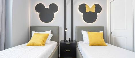 A twin room with two beds, each with white bedding and a yellow pillow. There are two nightstands with black drawers and a lamp on each. The walls feature a Mickey Mouse silhouette design.
