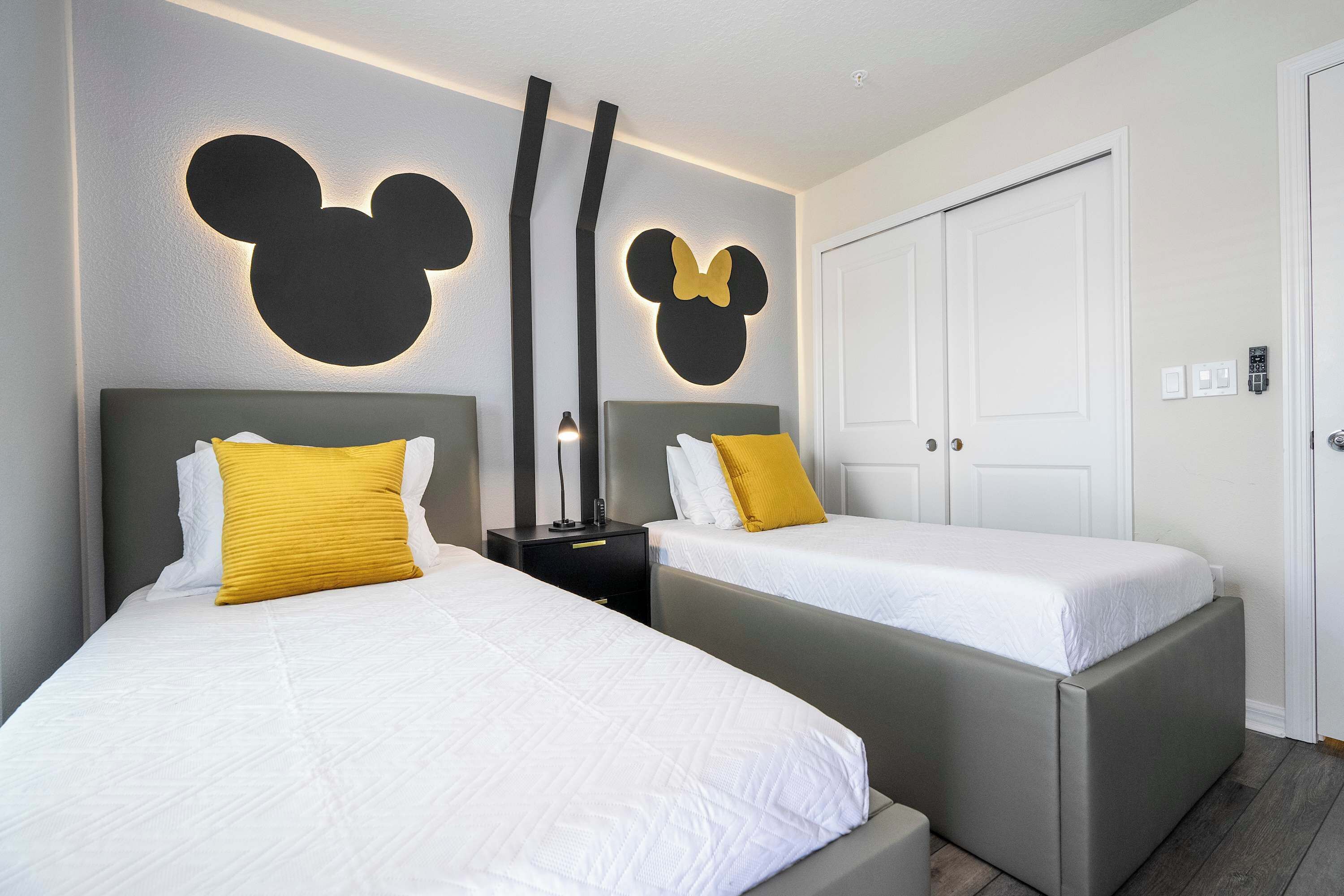 Ein Hotelzimmer mit zwei Betten, jeweils mit Kopfstütze und Kissen. An der Wand befinden sich Mickey-Mouse-Dekorationen, und die Tür ist mit einem Zahlenschloss versehen.